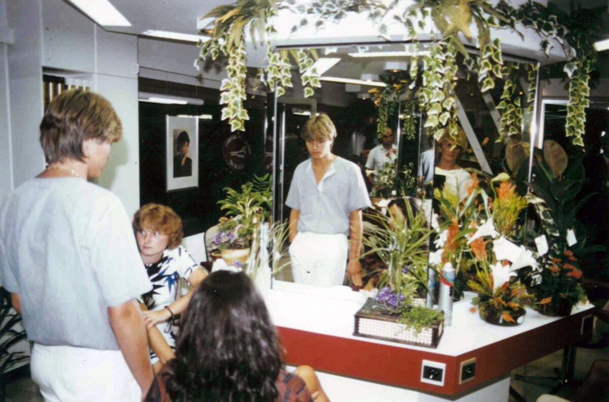 Drie jongeren en een vrouw zitten in een kapsalon, omringd door groene planten en bloemen, met spiegels op de achtergrond.