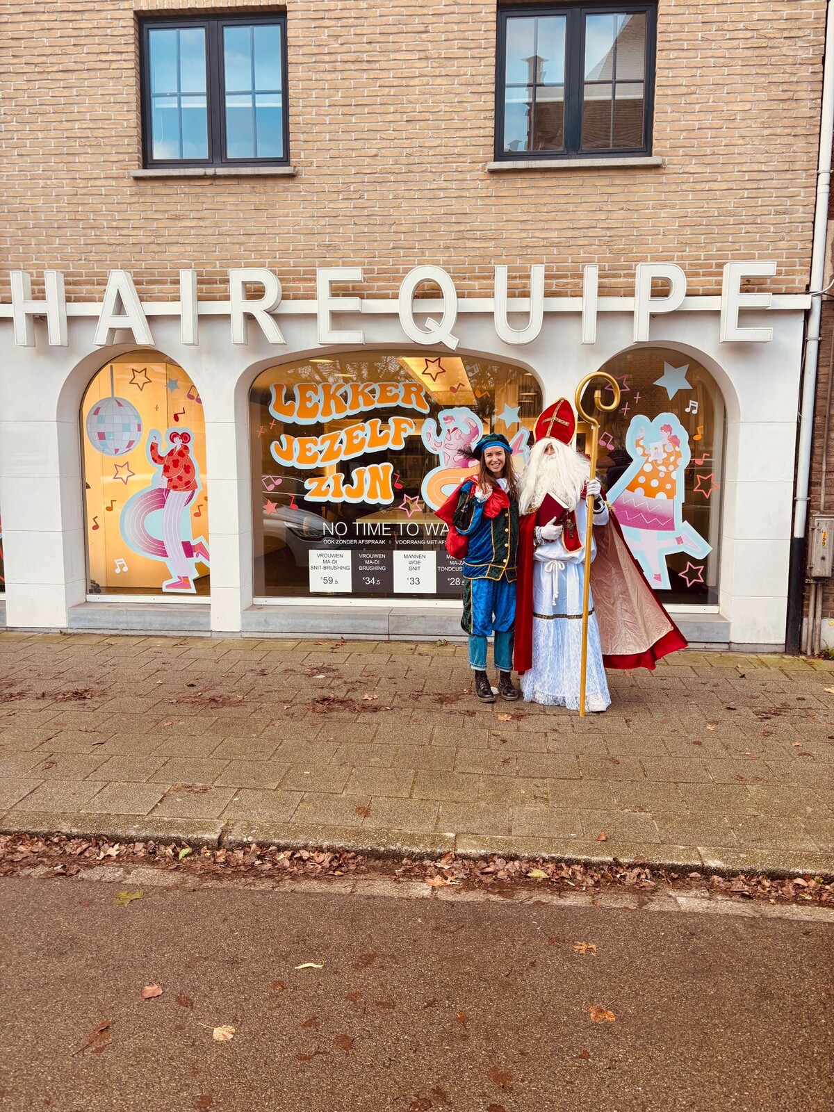 Een man en een vrouw staan voor een winkel met het teken "HAIREQUIPE". De man draagt een Sinterklaas-kostuum inclusief mijter en staf, terwijl de vrouw een kleurrijke culturele outfit draagt. Achter hen is een etalage met feestelijke decoraties en teksten.