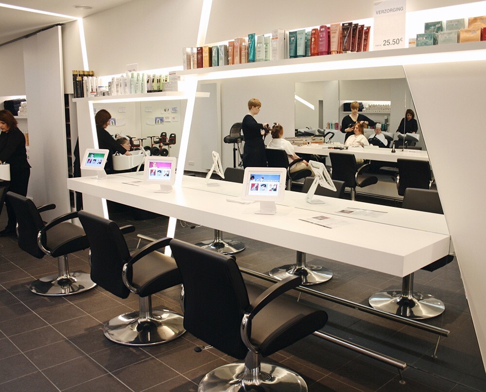 Een moderne kapsalon met een lange witte tafel en zwarte stoelen, waar klanten worden geknipt en verzorgt. Op de tafel staan tablets en er zijn verzorgingsproducten zichtbaar op een plank.