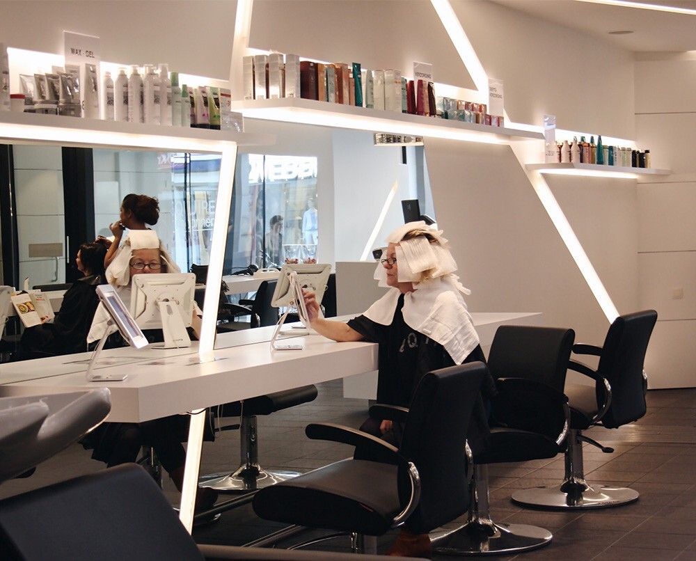 Een haarstylist in een moderne kapsalon kijkt in een spiegel terwijl ze haar haar laat doen, met andere klanten in de achtergrond en producten op de planken boven de werkplekken.