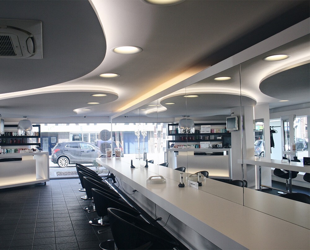 Modern interieur van een kapsalon met een lange witte balie, zwarte stylingstoelen, en een muur met spiegels en haarproducten.