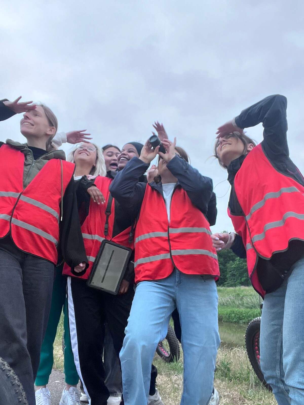 Groep jonge mensen in opvallende oranje vesten, lachend en handende richting de camera, met een groene omgeving op de achtergrond.