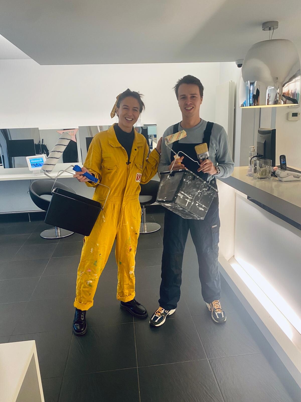 Twee jonge mensen staan in een moderne ruimte, beide gekleed in werkkleding en met verfbenodigdheden in hun handen. De vrouw draagt een gele overall met verfsporen, terwijl de man een zwarte overall en sneakers draagt. Ze glimlachen naar de camera.