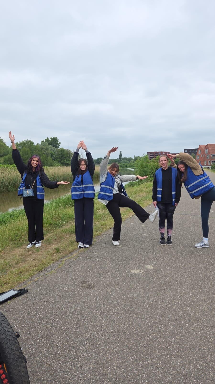 Vijf vrouwen in blauwe veiligheidsvesten poseren vrolijk langs een pad naast een waterloop, met een groene omgeving op de achtergrond en een lichtbewolkte lucht.