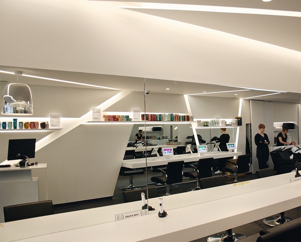 Moderne kapsalon met een strak interieur, spiegels en verzorgingsproducten op planken. Twee haarstylisten zijn bezig met klanten, en er zijn digitale schermen zichtbaar.