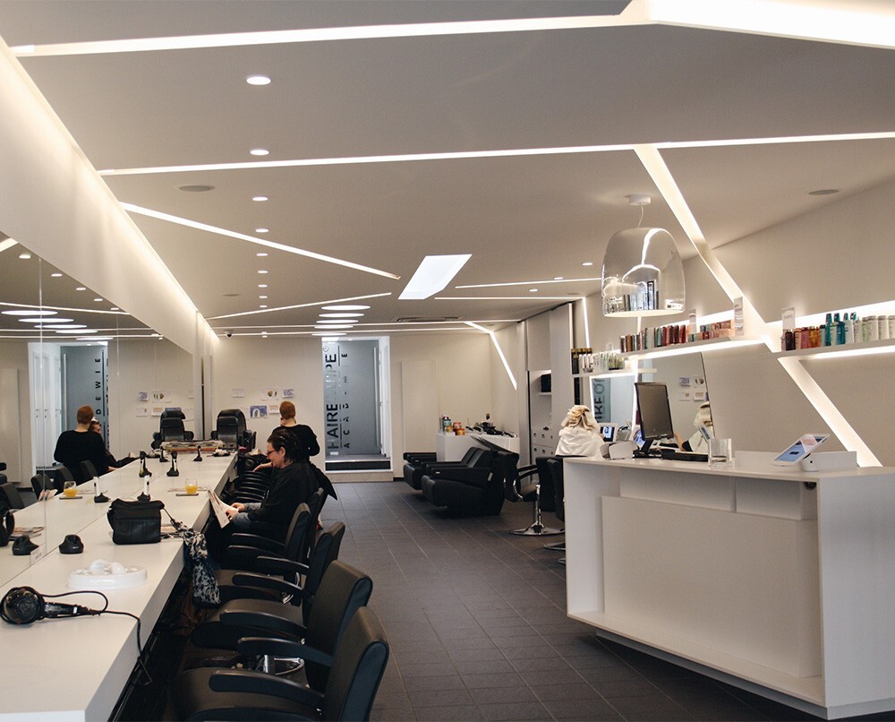 Interieur van een moderne kapsalon met een strak design, waaronder een lange witte balie, kappersstoelen en spiegels, verlicht door trendy verlichting.