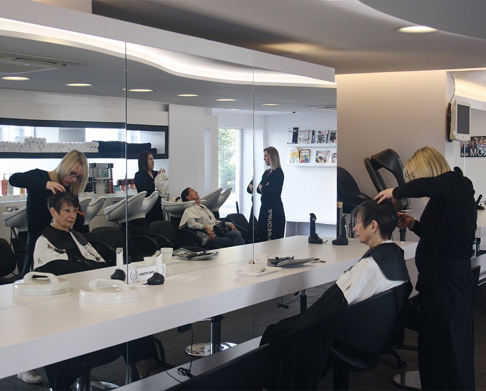 Een moderne kapsalon met meerdere klanten die een behandeling krijgen, en haarstylisten die werken aan de kapsels van de klanten. In de achtergrond zijn spiegels en verzorgingsproducten zichtbaar.