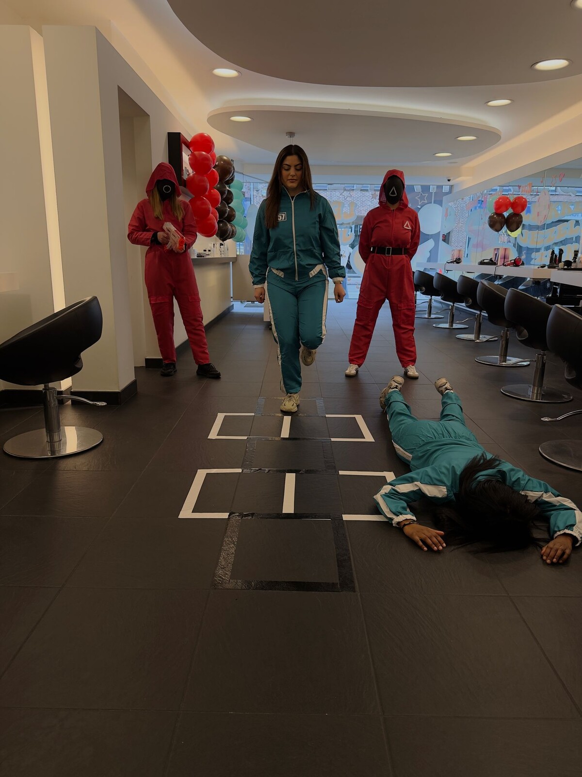 Drie personen in een salon, &eacute;&eacute;n vrouw in een turquoise outfit loopt over een speciaal gemarkeerd speelveld terwijl twee andere mensen in rode en zwarte kostuums toekijken; &eacute;&eacute;n persoon ligt op de grond. Ballonnen zijn zichtbaar op de achtergrond.