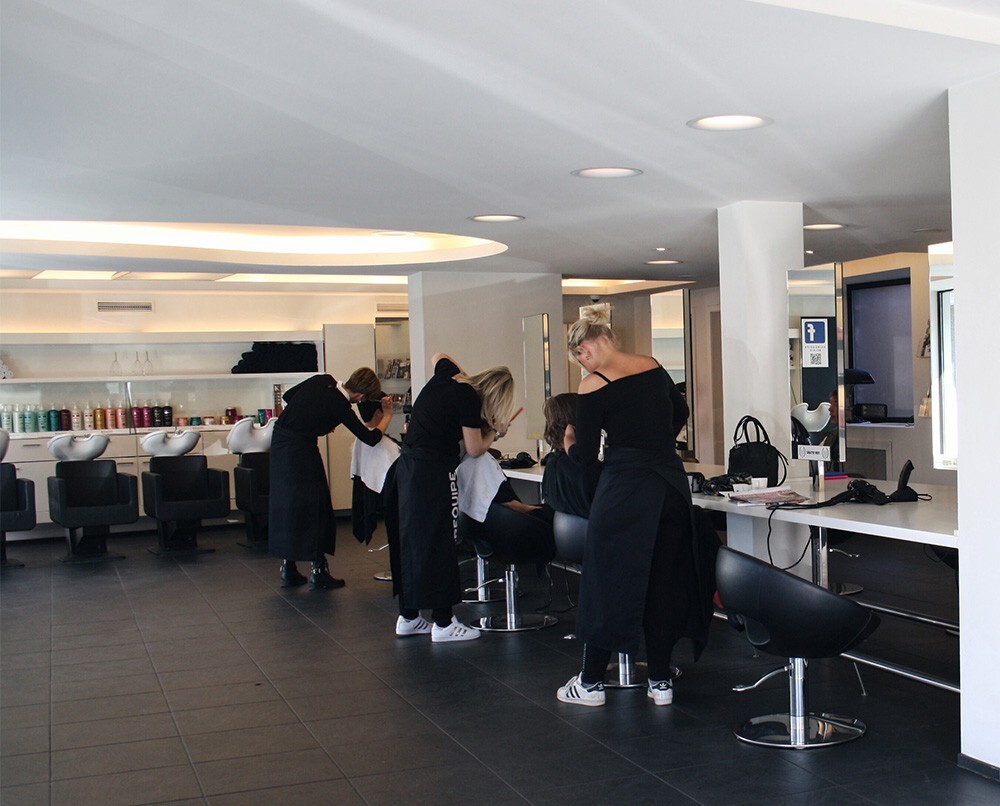 Een moderne kapsalon met drie stylisten die bezig zijn met hun klanten, terwijl ze gekleed zijn in zwarte schorten. Achter hen staan toiletten met haarkleurproducten zichtbaar op planken.